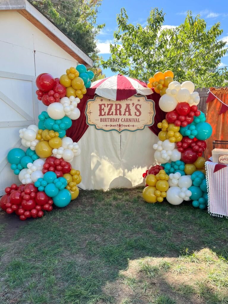 Carnival birthday balloon garland in red, teal, yellow and white around a name sign with a striped tent top