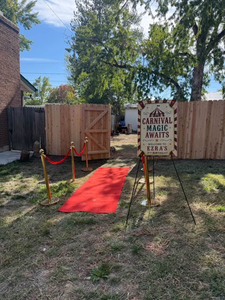 Red carpet entrance with rope stands and a sign on an easel in a backyard