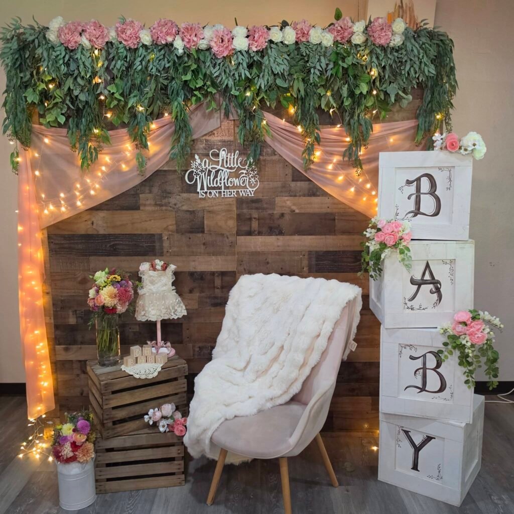 Rustic wood baby shower backdrop with a flower garland, string lights, BABY blocks and a white chair